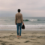 Мужчина стоит на берегу моря, держа в руках ботинки