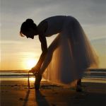 Балерина завязывает пуанты на фоне заката около моря